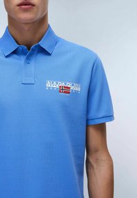 Chemise polo bleue en coton texturé avec patte de boutonnage à deux boutons. Affiche un logo sur la poitrine avec un détail du drapeau norvégien.
