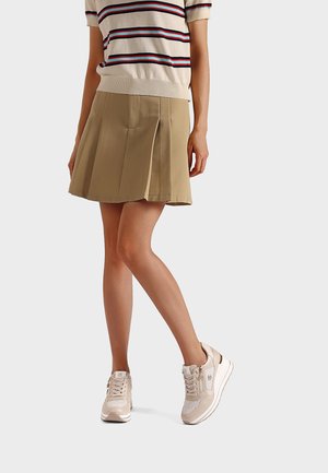 Donna con gonna beige plissettata, maglione a maniche corte a righe e sneakers beige con zeppa su sfondo neutro.