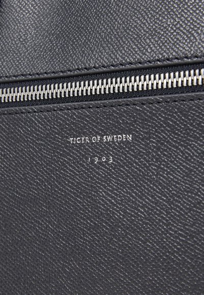 Cuir noir texturé avec un motif subtil, doté d'une fermeture éclair argentée et du logo en relief "Tiger of Sweden 1903".