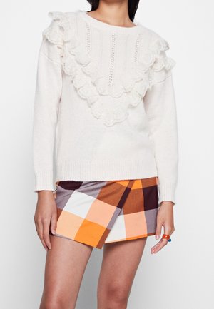 Maglione bianco lavorato a maglia con dettaglio in pizzo arricciato sul davanti abbinato a una gonna a portafoglio a quadri arancione, marrone e grigia.