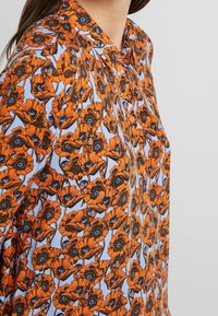 Chemise à imprimé floral orange avec un fond bleu clair, fermeture à boutons et col, ornée de contours noirs bien visibles des fleurs.
