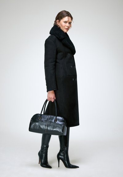 Femme portant un long manteau noir avec un col en fourrure, des bottes noires à talons hauts, tenant un grand sac à main en cuir noir sur un fond uni.