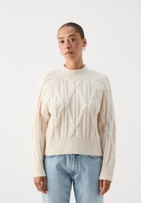 Πλεκτό πουλόβερ σε κρέμ απόχρωση με υφή διαμαντιών, ριμπ λαιμόκοψη και στρίφωμα, σε συνδυασμό με ανοιχτό μπλε τζιν. Χαλαρή εφαρμογή, μακριά μανίκια.