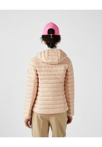 Giacca smanicata rosa chiaro con cappuccio, caratterizzata da sezioni trapuntate e una vestibilità aderente. Indossata con un cappellino rosa. Tessuto liscio con maniche lunghe.