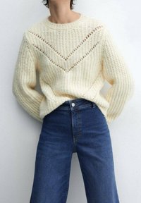 Roomkleurige dikke gebreide trui met chevronpatroon, gedragen met donkerblauwe jeans met hoge taille, persoon staat met handen achter de rug.