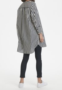 Vestido camisa de rayas en azul marino y blanco, con un ajuste ligeramente oversize, mangas largas y aberturas laterales; combinado con jeans oscuros y zapatos blancos.