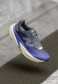 Γκρι και μοβ παπούτσι τρεξίματος New Balance με παχύ πάτο, που αιωρείται πάνω από άσφαλτο με λευκή γραμμή στο φόντο.