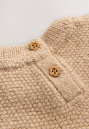 Beige strikket stof med et tekstureret mønster, der har to blomsterformede knapper i træ nær kraven og en stolpelukning nedenunder.