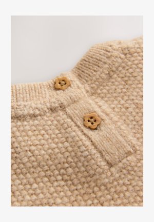 Beżowa dzianina o fakturowanym wzorze, z dwoma drewnianymi guzikami w kształcie kwiatów przy kołnierzu oraz detalem pliski pod spodem.
