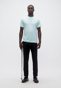 Άνδρας που στέκεται κρατώντας ένα κλαμπ γκολφ κάθετα, φορώντας ανοιχτό μπλε μπλουζάκι, μαύρο παντελόνι και μαύρα αθλητικά παπούτσια σε απλό λευκό φόντο.