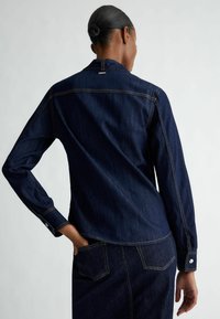 Camicia in denim di colore blu scuro con maniche lunghe, polsini a bottoni e cuciture visibili. Presenta un yoke posteriore e due tasche sul retro.