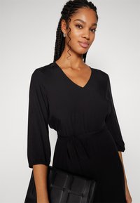 Robe noire avec un décolleté en V et des manches 3/4, dotée d'une ceinture à nouer. Le modèle tient un sac à main noir texturé. Tissu lisse et léger.