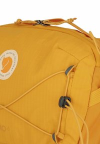 Zaino giallo con una finitura testurizzata, dotato di un'etichetta con logo, corde elastiche e accessori grigi. Dimensioni ridotte, adatto per il trekking.