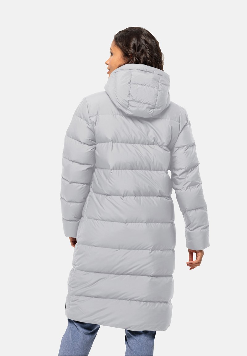 Donna che indossa un lungo cappotto imbottito grigio chiaro con cappuccio, in piedi di spalle contro uno sfondo bianco semplice.
