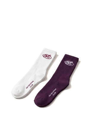 Une chaussette blanche et une violette de sport avec une texture côtelée, chacune arborant le logo "OVC" près du poignet et le texte "ON VACATION CLUB" sur le pied.