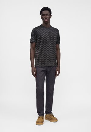 Homme debout portant un t-shirt noir à motif zigzag, jean foncé et chaussures décontractées marron clair sur un fond clair uni.