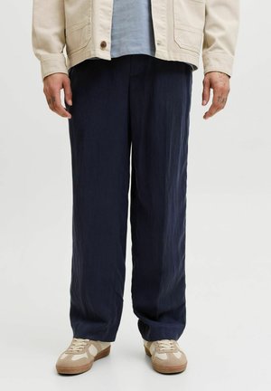 Homme portant un pantalon ample bleu foncé, des baskets beiges, une chemise bleu clair et une veste beige, debout devant un fond clair uni.