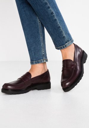 Person trägt dunkelbraune Leder-Loafers und blaue Jeans und steht auf einem hellen Boden mit einem leicht angehobenen Fuß.