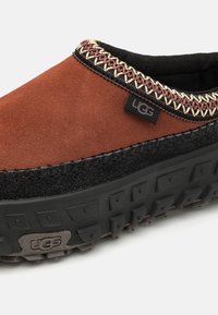 Barna és fekete UGG slip-on cipő vastag fekete talppal, díszvarrással a nyílás körül, és UGG logós címkével az oldalon.
