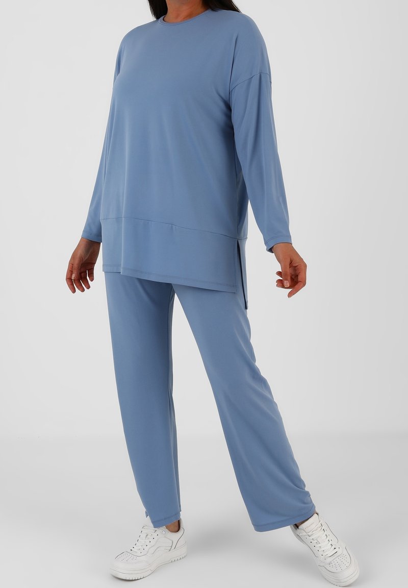 Femme portant un haut bleu à manches longues ample et un pantalon assorti avec des baskets blanches, debout devant un fond clair uni.