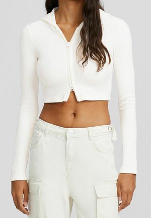 Vrouw draagt een witte cropped geribbelde zip-up hoodie en gebroken witte cargobroek met klepzakken, getoond van nek tot heupen.