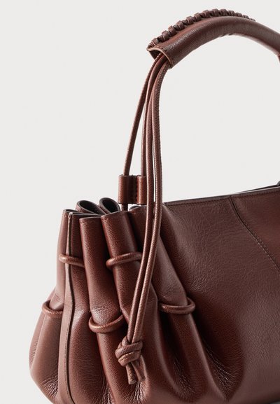 Καφέ δερμάτινη γυναικεία τσάντα με μαζεμένες πλευρές, διακοσμητικά λουράκια με κόμπους και τυλιγμένη λαβή σε απλό ανοιχτό φόντο.