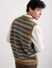 Uomo con capelli ricci e scuri che guarda oltre la spalla, indossando un gilet lavorato a maglia sopra una camicia a maniche lunghe crema, su uno sfondo semplice.