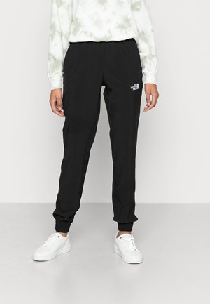 Osoba nosząca czarne spodnie dresowe The North Face, białe sneakersy oraz jasną bluzkę z długim rękawem i subtelnym wzorem, stojąca na jednolitej podłodze.