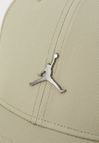 Beige cap med tekstureret stof, har et metal Jumpman-logo på forsiden, syede sømme og en buet skygge.