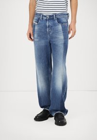 Wide-leg jeans in medium blue denim met vervaagde plekken, vijf zakken en een knoopsluiting, gecombineerd met zwarte schoenen.