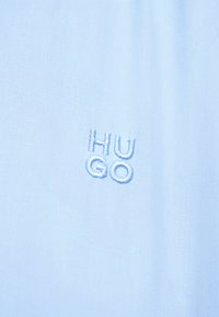 Ανοιχτό μπλε ύφασμα με το κέντημα "HUGO" σε ελαφρώς πιο σκούρα μπλε κλωστή, διατεταγμένο σε δύο σειρές, δύο γράμματα σε κάθε σειρά.