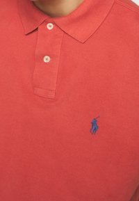 Detailní záběr na červenou polo tričko se dvěma bílými knoflíky a malým modrým vyšitým logem hráče póla na hrudi.