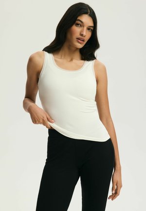 Kobieta o długich ciemnych włosach, ubrana w biały bezrękawnik i czarne spodnie, trzymająca jedną ręką krawędź swojego topu.