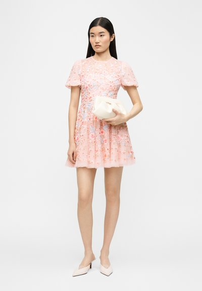 Νεαρή γυναίκα με μακριά μαύρα μαλλιά φοράει κοντό φόρεμα ροζ με κεντήματα λουλουδιών, κρατάει λευκή μικρή τσάντα και φοράει ανοιχτό ροζ τακούνια.