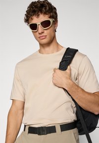 Beige bomull t-skjorte, svart belte og mørk stropp ryggsekk. Grå solbriller med brune linser. Brukt med khaki bukser.