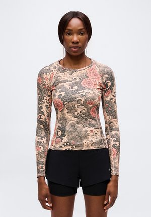 Kvinde iført bluse med lange ærmer med blomster- og bølgemønster med Champion-logoer og sorte shorts, der står mod en ensartet lys baggrund.