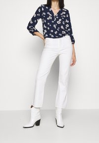 Námořnická květinová košile na knoflíky s dlouhými rukávy, spárovaná s bílými kalhotami s vysokým pasem a rovnými nohavicemi a bílými botami na podpatku kolem kotníku.