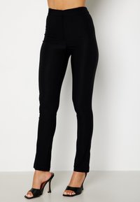 Pantalon noir ajusté avec une texture lisse et une taille haute, présentant une coupe droite et des coutures latérales discrètes, associé à des talons noirs.