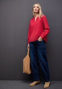 Camisa roja de cuello con mangas largas, combinada con jeans de pierna ancha azules y botas de tobillo de ante beige. Sosteniendo un bolso tote de ante beige.