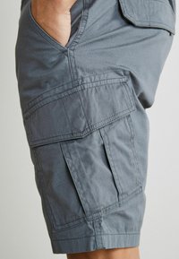 Szürke cargo shorts, könnyű anyagból készült, oldalsó zsebekkel, varrott díszítéssel és laza szabással. A textúra simának és tartósnak tűnik.