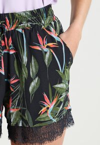 Žena v černých květovaných šortkách s krajkovým lemem, ruka v kapse, a světle fialovém topu.