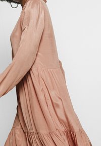 Donna che indossa un vestito lungo, a maniche lunghe, a strati di colore rosa polveroso, realizzato in un tessuto morbido e leggermente lucido, su uno sfondo bianco semplice.