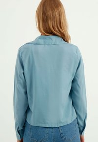 Lichtblauwe, lichte blouse met lange mouwen en kraag, gladde textuur en met knopen afgewerkte manchetten; weergegeven vanuit de achterkant.