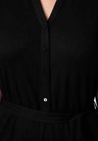 Robe noire texturée avec un devant à boutons, décolleté en V et une ceinture nouée à la taille. Présente un léger éclat et est fabriquée en tissu léger.