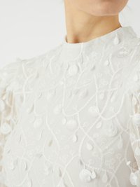 Bílá vyšívaná látka s nepravidelnými květinovými vzory, nabírané rukávy a vysoký límec. Textura je jemná a průsvitná.