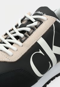 Svart og beige sneakers med en glatt overflate, hvite snørebånd og en stor hvit "CK" logo på siden. Har teksturerte materialer og kontrastaksenter.