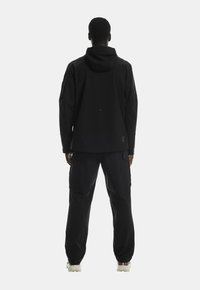 Černá nepromokavá bunda a kalhoty, se kapucí, volným střihem a cargo kapsami, doplněné světlou obuví.