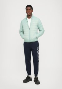 Jachetă puffer de culoare verde deschis, cu cusături orizontale, închidere cu fermoar și guler înalt, asortată cu pantaloni de trening bleumarin cu imprimeu alb logo.