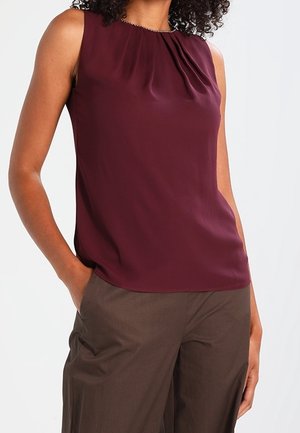 Femme portant un chemisier sans manches bordeaux avec des plis au niveau de l'encolure et un pantalon marron ample, debout, une main dans la poche.