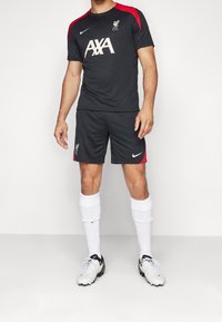 Svart Nike fotbollskit med röd accent, logga på bröstet, matchande shorts och vita strumpor. Skorna är svarta och vita.